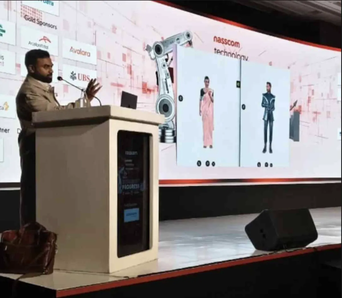 A speaker on stage at the NASSCOM Tech Conference with virtual characters displayed on the wall behind